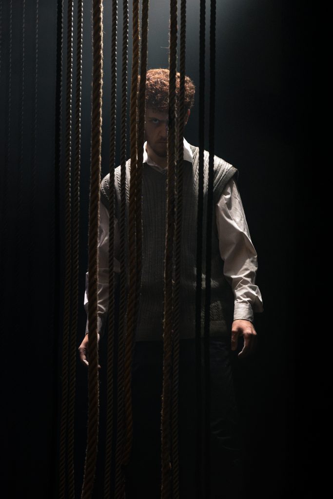 An Actor lurks behind some ropes hanging in the space, the lighting is atmospheric and spooky. His features are partially hidden.