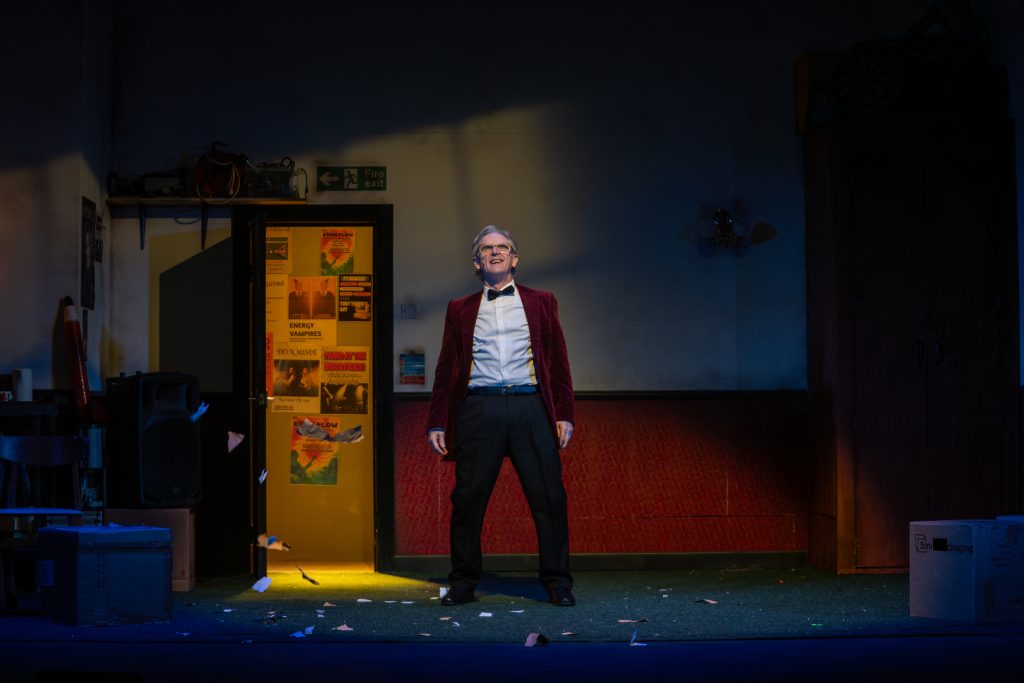 Harry (Andrew Schofield) Stands in a red blazer, in the middle of the run down dressing room in his social club